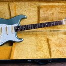 Elektrokytara s modrozeleným trupem a bílým pickguardem v černém kufru s žlutým polstrováním. Na hlavě hmatníku je viditelné logo Fender a nápis STRA…