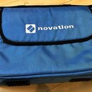 Modrá textilní brašna s černými zipy a okraji. Uprostřed horní klopy je bílý nápis novation s doprovodným logem v čtverci. Povrch materiálu je v dobr…
