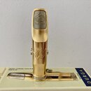 Zlatý kovový mouthpiece s šedou náustníkem, na kterém je vyrytý ornamentální symbol. Na těle mouthpieceu je vyryto logo ve tvaru stylizovaného písmen…
