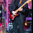 Fotografie zachycuje hudebníka na pódiu s elektrickou kytarou typu Stratocaster v červené barvě s bílým pickguardem. Kytara má javorový hmatník a je…