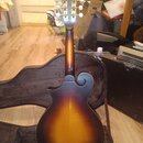 Zadní pohled na mandolínu s tmavě hnědým a žlutým přechodovým lakem (sunburst). Nástroj má vyřezávané průduchy v těle a bílé ladičky. Na hlavě nástro…