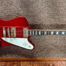 Červená elektrická kytara s atypickým asymetrickým tvarem těla a bílým pickguardem. Na hlavě hmatníku je viditelné logo Epiphone. Nástroj má dva sním…