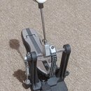 Černý bicí pedál s kovovými mechanismy a bílou gumovou koncovkou na šlapadle. Na kovové části je viditelný nápis "TAMA". Povrch černého plastu vykazu…