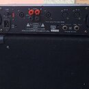 Zadní strana černého rackového zesilovače s nápisem The t.amp TA1400 MKII. Zařízení má napájecí vstup AC 220-240V, dva červené konektory OUT A a OUT…