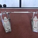 Dvojpedál na bicí buben z kovového materiálu v stříbrné barvě. Na obou pedálech je umístěn nápis Pearl a čtyři červené gumové body. Povrch vykazuje z…