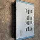 Stříbrno-modrý kovový box s ventilačními otvory ve tvaru tří kosočtverců na horní straně. Boční stěna obsahuje řadu modrých konektorů. Povrch vykazuj…