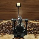 Fotografie zobrazuje basový pedál v černé barvě s kovovými mechanismy a řetězem. Pedál má černý šlapadlo s bílým polstrováním v horní části. Na zaříz…