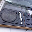 Gramofon v dřevěném šasi s černým horním panelem a průhledným plastovým krytem. Na ovládacím panelu je nápis NZEC 180 STEREO a zelené tlačítko s nápi…
