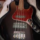 Černá elektrická basová kytara v otevřeném obalu. Nástroj má červený vzorovaný pickguard a na snímači je nápis BASSLINES. Povrch kytary vykazuje drob…