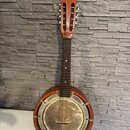 Akustický banjo s hnědým dřevěným tělem a krkem. Instrument má kovový, stříbrný rezonanční povrch s viditelnými šmouhama a oxidací. Na hmatníku jsou…