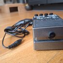 Kovový efekt pedál v šedé barvě s nápisem Reverb a logem BOSS. Na horní části jsou vidět čtyři konektory s popisky OUTPUT, OUTPUT 2 a INPUT. K pedálu…