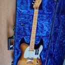 Elektrická kytara s hnědým průsvitným lakem a bílým pickguardem v modrém polstrovaném kufru. Na hlavě hmatníku je nápis Fender. Tělo kytary má zlaté…