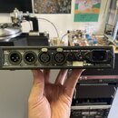 Zadní strana černého elektronického zařízení s kovovým šasi. Jsou zde čtyři konektory XLR, jeden konektor typu DIN a napájecí vstup. Na povrchu jsou…