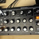 Černý analogový syntetizátor s nápisem MINITAUR analog bass synthesizer a logem Moog v levém dolním rohu. Zařízení má kovové tělo s mnoha stříbrnými…