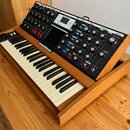 Klávesový syntetizátor v dřevěném rámu s černým ovládacím panelem. Na panelu jsou vidět nápisy "NOVATION", "MIXER", "FILTERS", "ENVELOPES" a "OUTPUT"…