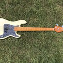 Elektrická basová kytara s krémovým korpusem a hnědým javorovým hmatníkem. Na korpusu je šedý pickguard s texturou mramoru a stříbrný most. Nástroj n…
