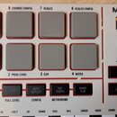 Detailní pohled na ovládací panel bílého MIDI kontroléru s nápisem MPK mini. V horní části je osm šedých čtvercových padů v červeném rámu s popisky 5…