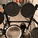 Elektronická bicí souprava s černým rámem a gumovými pady. Na bílém povrchu basového bubnu je nápis Roland a logo firmy Remo s textem REMO. Povrchy p…
