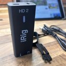 Černý obdélníkový modul s nápisy HD 2 a iRig. Zařízení má matný povrch, v horní části otvor pro kabel a na boku malý symbol kytary. Vedle modulu leží…