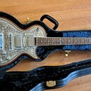 Elektrická kytara s kovovým povrchem zdobeným rytinami květin a listů, umístěná v černém tvrdém kufru. Na těle jsou dva humbuckery a čtyři ovládací k…