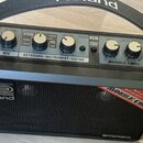 Černý přenosný stereo zesilovač s rukojetí. Na horním ovládacím panelu jsou knofíky a nápisy VOLUME, STEREO INPUT KEYBOARD/INSTRUMENT (NORMAL, A-GUIT…