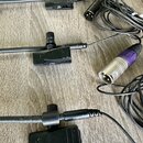 Tři černé mikrofony s klipy na oblečení a připojenými kabely. Na kovových konektorech je vidět nápis XLR. Povrchy plastových částí vykazují mírné zná…