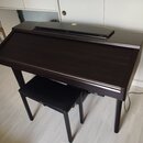 Tmavně hnědý digitální piano v podobě konzole s přívukovou stoličkou čalenou černým materiálem. Na stoličce je vidět nápis Yamaha. Povrch nástroje je…