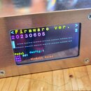 Detailní pohled na malý digitální displej osazený v kovovém, stříbrném šasi s viditelnými šrouby. Na displeji je zobrazen text v různých barvách: <Fi…