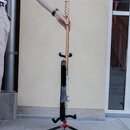 Elektrická kytara černé barvy umístěná na černém trojnožném stojanu. Nástroj má světlý hmatník a dřevěnou hlavu s kovovými ladičkami. Na fotce nejsou…