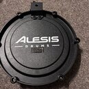 Černý plastový pad elektronických bubnů s kulatým tvarem. Uprostřed je bílý nápis ALESIS DRUMS a pod ním malá stříbrná štítek s technickými údaji, lo…