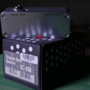 Kovový efekt pedál stříbrné barvy položený na černé krabici. Na horní části pedálu jsou nápisy INPUT DC 9V a BATTERY INSIDE, na spodní hraně je obrác…