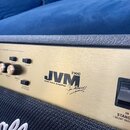 Detailní záběr na horní ovládací panel kytarového komba v zlaté barvě. Na panelu je černý nápis JVM 210C a text 100W Valve Amplifier spolu s podpisem…