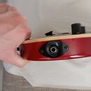 Detailní pohled na spodní hranu červené elektrické kytary. Je zde vidět vstupní jack s černým plastovým krytem přišroubovaným dvěma šroubky. Na horní…