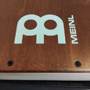 Detail dřevěného nástroje v hnědé barvě s viditelnou strukturou dřeva. Na povrchu je bílý nápis MEINL a vedle něj velké bílé logo ve tvaru stylizovan…