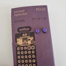 Fialové papírové obalení s nápisy pocket operator, PO-20, arcade a japonskými znaky アーケード. V obalu je umístěn kompaktní syntezátor s hnědým předním p…
