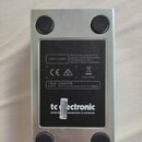 Fotografie zachycuje spodní stranu stříbrného kovového efektpedálu s čtyřmi černými gumovými nožičkami. Na černé štítku je uveden název tc electronic…
