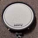 Kruhová elektronická bubenová pad v černé barvě s bílou úderovou plochou. Na spodní části plochy je černý nápis ALESIS DRUMS. Pad má černý obrubník a…
