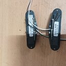 Dvě elektrické snímače kytary s černými plastovými kryty a kovovými pružinami, umístěné na černých montážních deskách. Na obou snímačích jsou bílé pa…