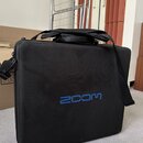 Černý textilní obal s rukojetí a ramenním popruhem. Na přední straně je modrý nápis ZOOM. Povrch materiálu vykazuje lehké znečištění a opotřebení.