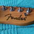 Detail hlavy elektrické kytary z přírodního dřeva s bílou maticí. Na dřevě je černý nápis Fender®. Jsou vidět šest kovových ladících kol. Povrch dřev…