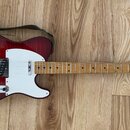 Elektrická kytara s červeným lakem s viditnou strukturou dřeva, bílým pickguardem a hnedým hmatníkem. Na hlavě je logo "Squier" a nápis "TELECASTER".…