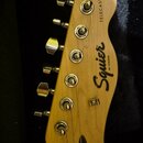 Fotografie zachycuje hlavu elektrické kytary z přírodního světlého dřeva. Na povrchu je černý nápis Squier BY FENDER a svislý nápis TELECASTER®. Jsou…