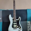 Bílá elektrická kytara typu Stratocaster s černým pickguardem a hnědým hmatníkem. Na hlavě je viditelné logo Fender. Nástroj má tři snímače, tři bílé…
