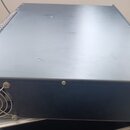 Černý kovový přístroj v rackovém provedení. Na zadní straně je viditelný chladicí ventilátor s kovovou mřížkou. Horní kryt je plochý a v levém horním…