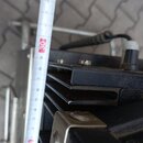 Detailní pohled na černý kovový přístroj s chladicími žebry a několika ovládacími knoflíky. Vedle přístroje je přiložen bílý skladací metr, který uka…