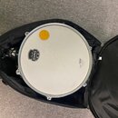 Bílý basový buben v černém polstrovaném obalu. Na blaně je vidět logo MAPEX uvnitř černého štítu a nápis LT s malou korunkou a textem "DRUM HEADS MAD…