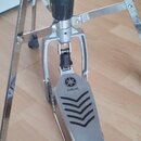Detailní pohled na kovový pedál bicího bubnu v stříbrné barvě. Na plošince je viditelné logo a nápis YAMAHA s černými dekorativními vlnovkami. Konstr…