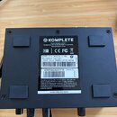 Zadní strana černého audio rozhraní s nápisem KOMPLETE. Na štítku je uveden model Komplete Audio 1, text Designed by NATIVE INSTRUMENTS in Germany. M…
