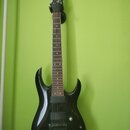 Černá elektrická kytara s tmavým hmatníkem, zavěšená na zelené stěně. Na hlavě nástroje je bílý nápis Ibanez. Kytara má dva humbuckery, jeden ovládac…