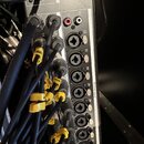 Detailní pohled na zadní panel audio zařízení s řadou vstupů. Jsou zde vidět konektory XLR a jack, do kterých jsou zapojeny černé kabely s žlutými oz…